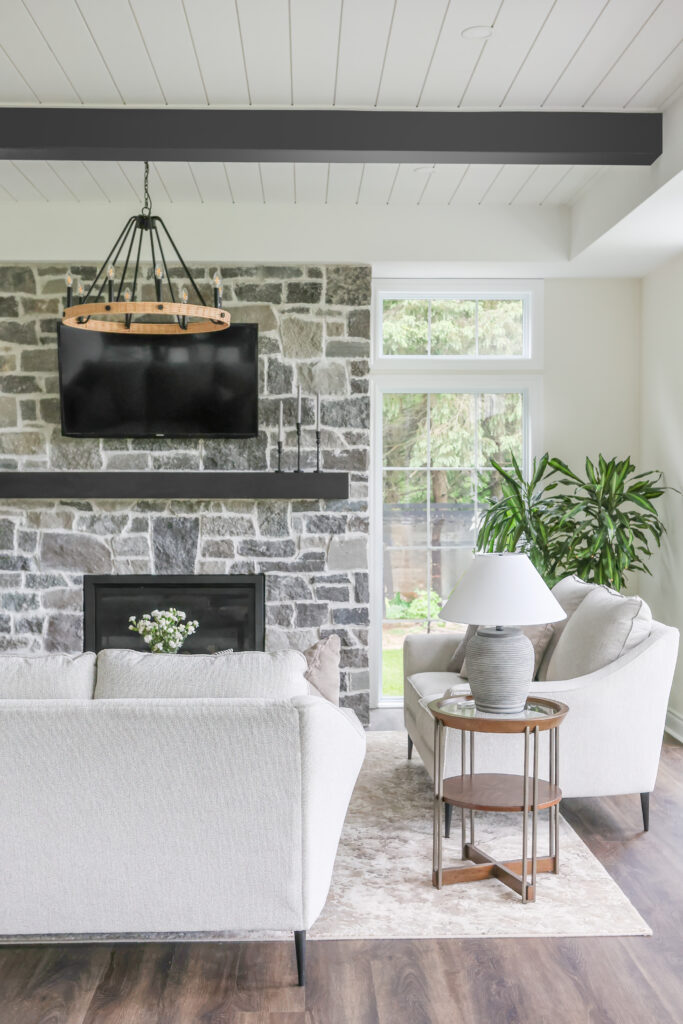 A modern living room with a stone accent wall, wall-mounted TV, fireplace, light sofas, side table with a lamp, potted plant, and large windows letting in natural light.