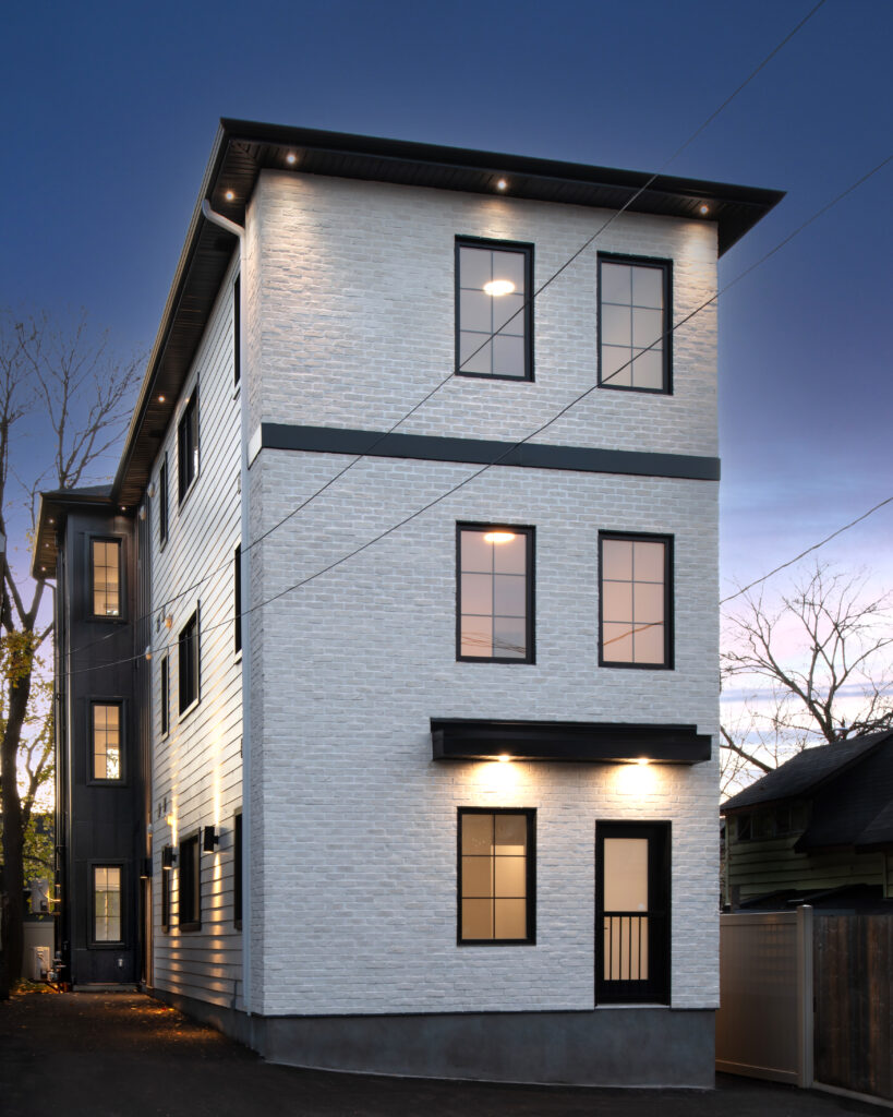 A modern three-story building with white brick walls, black trim around the windows, and exterior lights on, photographed during dusk. There are bare trees and neighboring houses visible in the background.