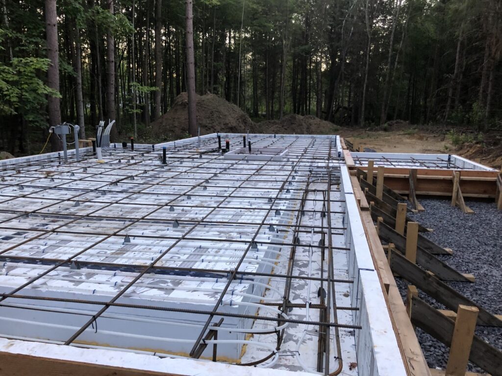 A construction site shows a foundation prepared for concrete pouring, with rebar grid and pipes in place, surrounded by trees and dirt piles in the background.