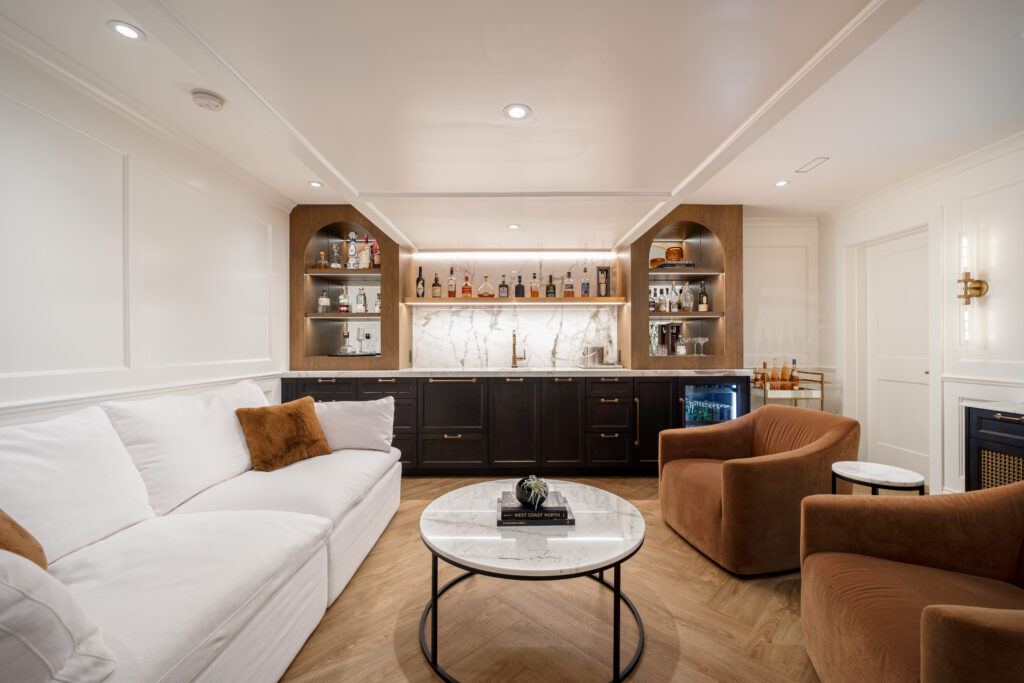 A modern home bar and lounge with a white sofa, two brown armchairs, round coffee table, wooden floors, and a backlit marble bar with shelves of bottles and glasses.