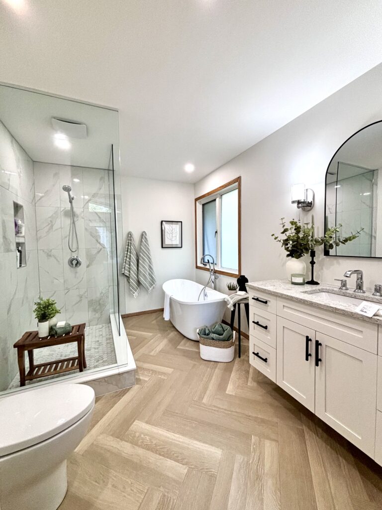 Modern bathroom with a glass shower, freestanding bathtub, white vanity with a marble countertop, oval mirror, wood herringbone floor, potted plants, striped towels, and natural light from a window.