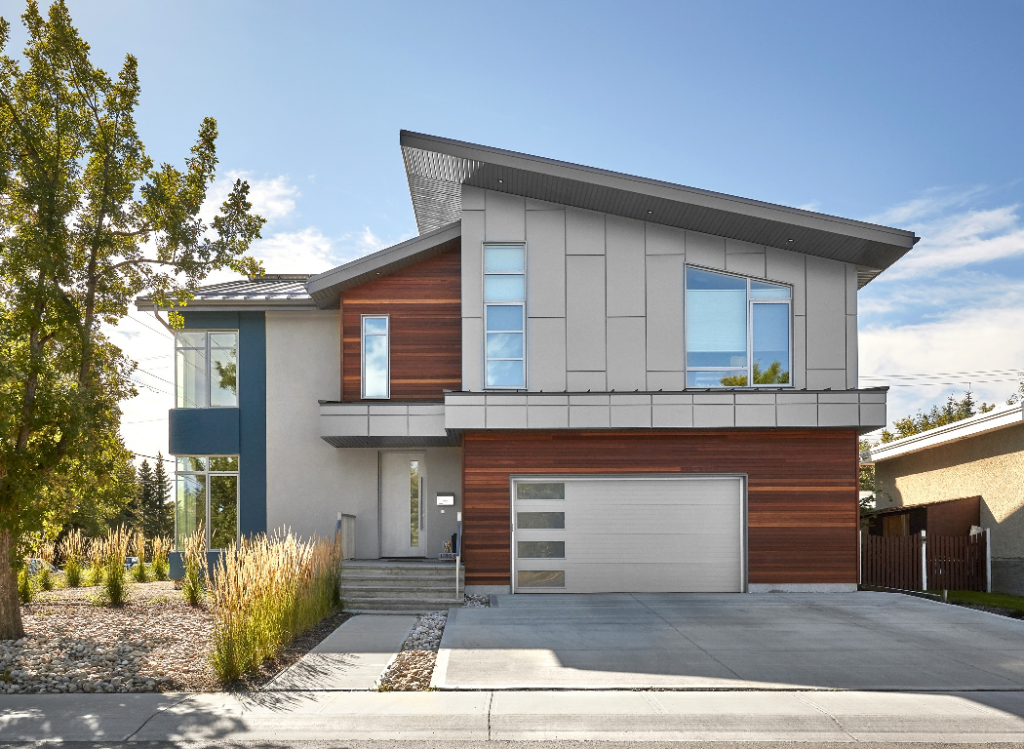 A modern two-story house with a sloped roof, wood and gray panel exterior, large windows, and a double garage. The home is surrounded by landscaping with rocks, grass, and a tree on the left.