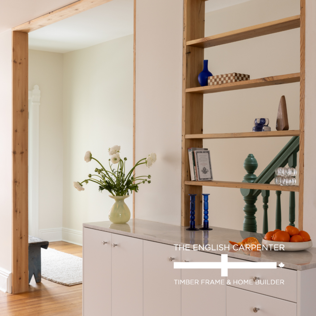 A modern home interior with white cabinets, a built-in wooden shelf holding vases and decor, a vase of flowers on the counter, green staircase spindles, and a bowl of oranges. Logo text: The English Carpenter Timber Frame & Home Builder.