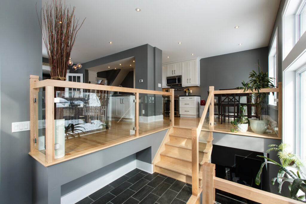 Bright, modern split-level home interior with wooden stairs, glass railings, gray walls, white kitchen cabinets, large windows, and potted plants; airy and contemporary design with natural light.