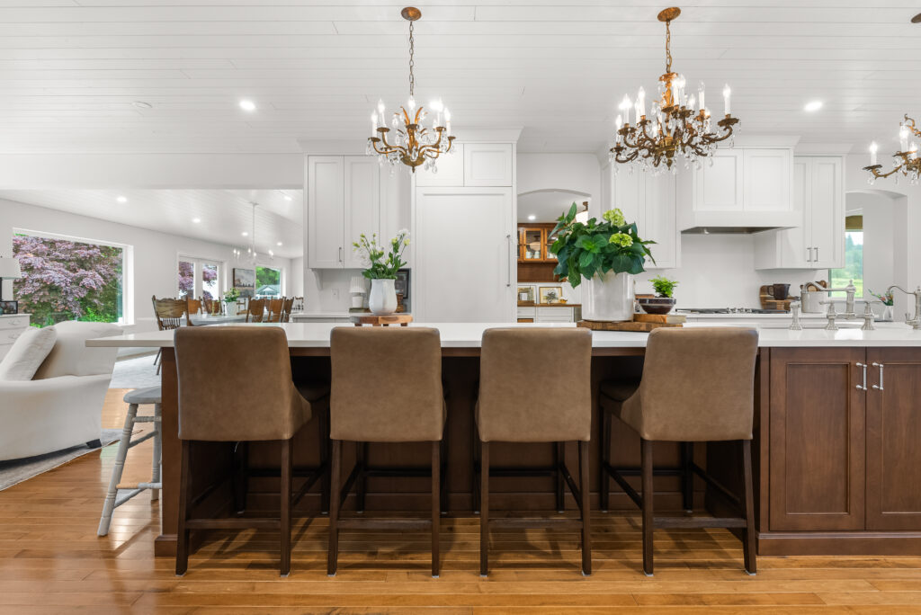 Spacious, modern kitchen with a large island, brown chairs, white cabinetry, two chandeliers, plants, and hardwood floors. Open layout extends to dining and living areas with ample natural light.