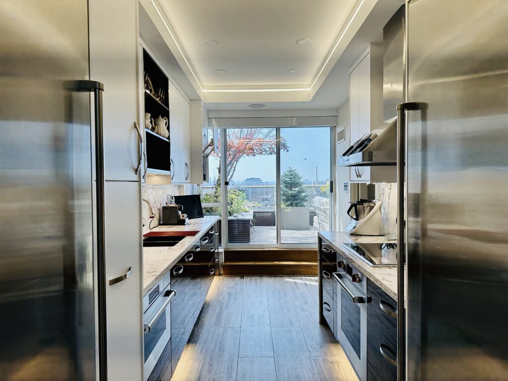 Modern galley kitchen with stainless steel appliances, white cabinets, and wood flooring, leading to a bright balcony with glass doors and a view of trees and a cityscape outside.