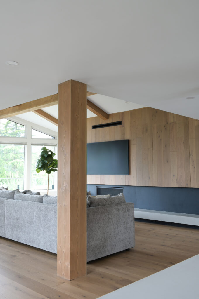 A modern living room with light wood floors, a wood-paneled accent wall with a mounted TV, large windows, a gray sectional sofa, and a tall green plant by the window. A wooden support beam is centered in the image.