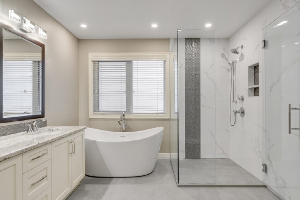 Modern bathroom with a freestanding white bathtub, large glass-enclosed shower, beige walls, double-sink vanity with light countertops, and windows with white blinds.