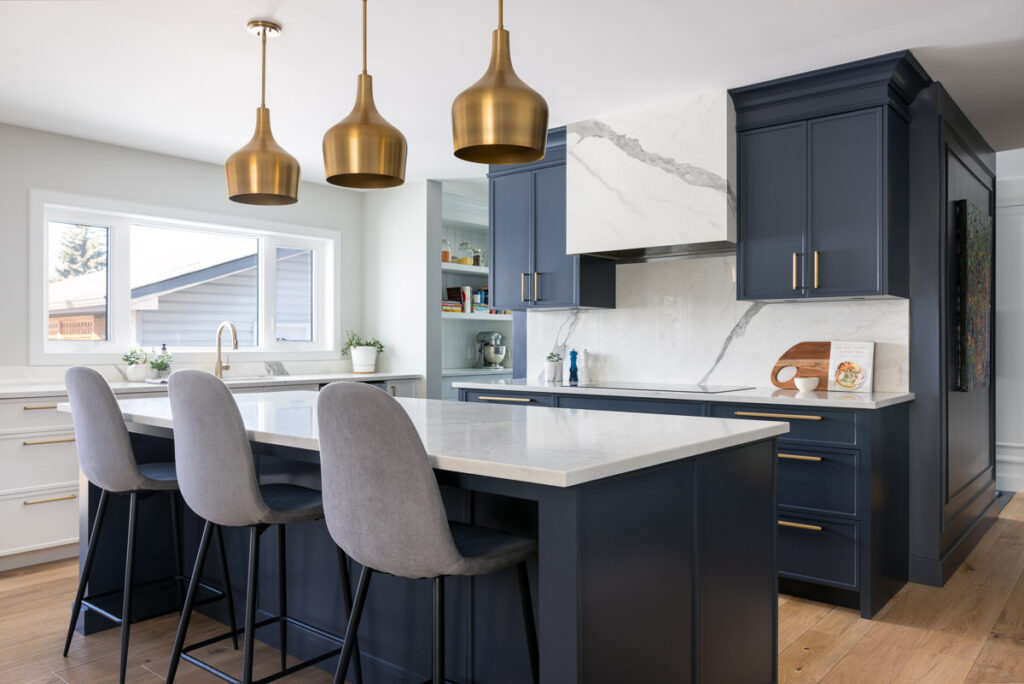 Modern kitchen with navy blue cabinets, white countertops, and a large island with three gray upholstered chairs. Three gold pendant lights hang above the island. Light wood flooring and a marble backsplash are also visible.