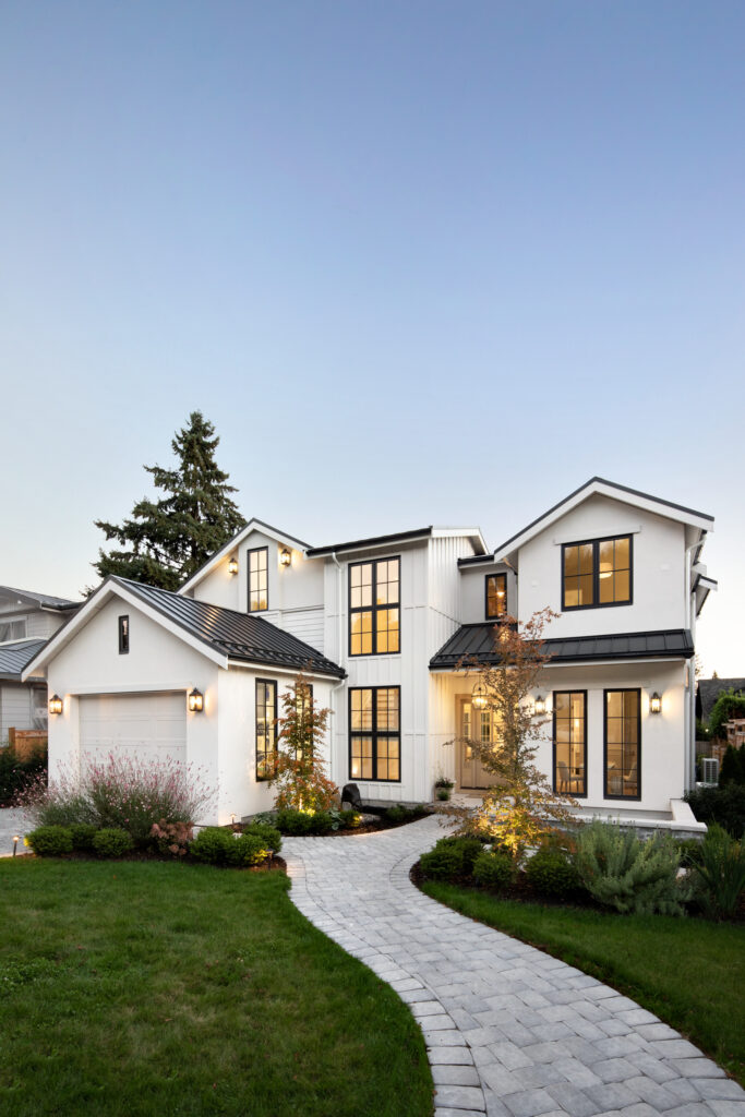 Modern two-story white house with black trim and large windows, featuring a well-kept lawn, landscaped bushes, and a curved stone pathway leading to the front entrance at dusk.