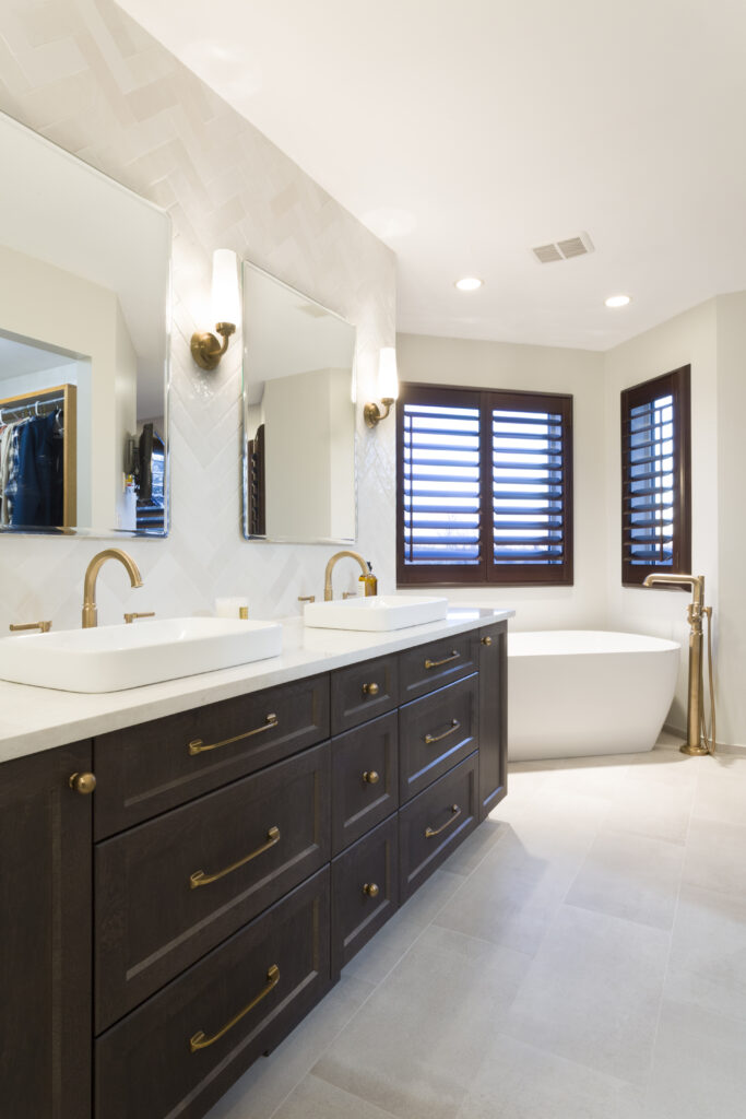 Modern bathroom with double dark wood vanity, gold fixtures, vessel sinks, two large mirrors, wall sconces, a freestanding white bathtub, and large windows with wooden shutters letting in natural light.