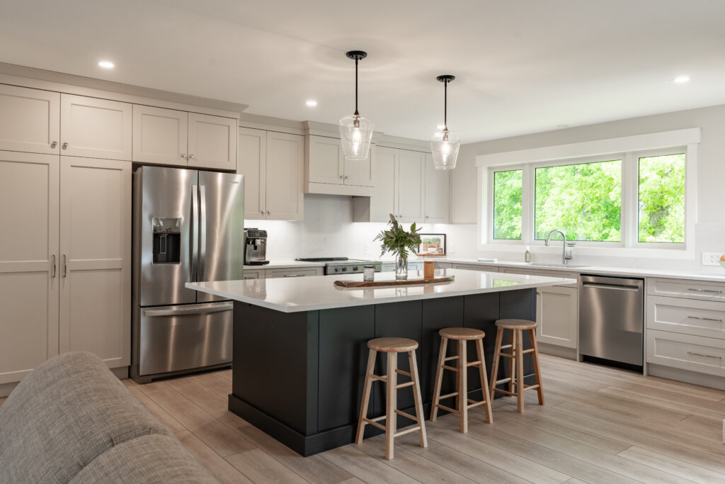 Modern kitchen with light gray cabinets, stainless steel appliances, a large dark island with a white countertop, three wooden stools, pendant lights, and a window overlooking greenery.