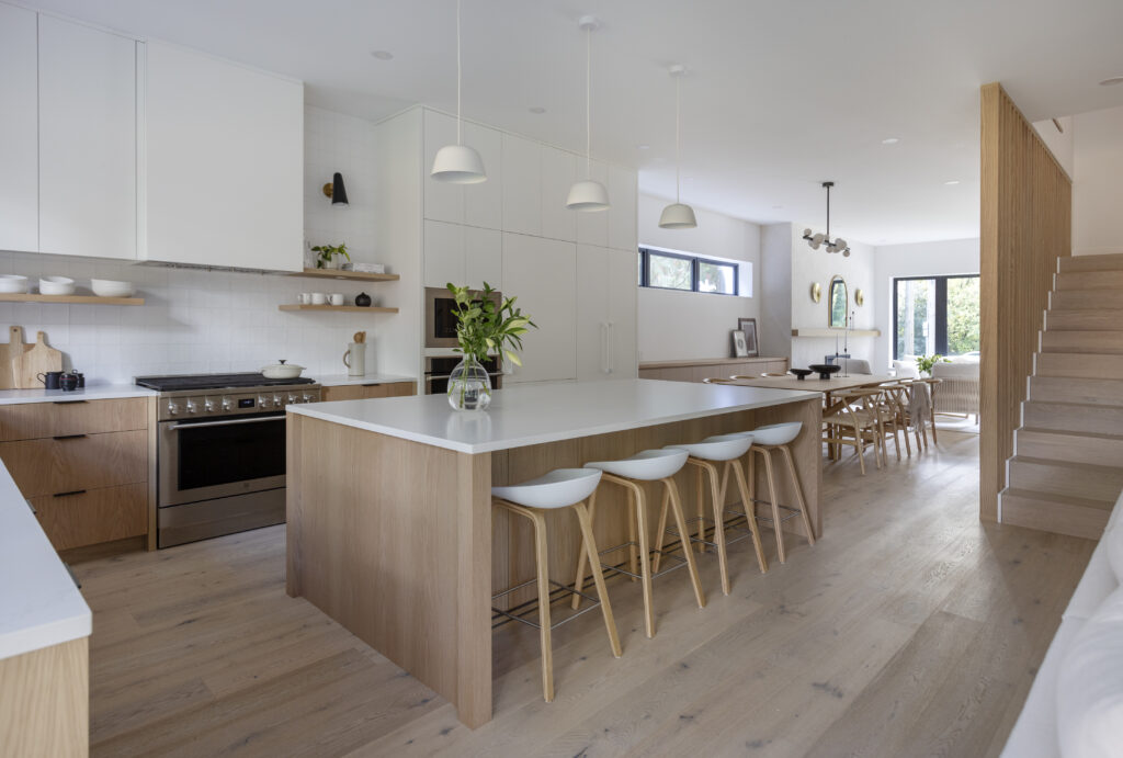 A modern, open-concept kitchen with light wood floors, a large island with white countertops and barstools, white cabinets, stainless steel appliances, pendant lights, and a dining area in the background.