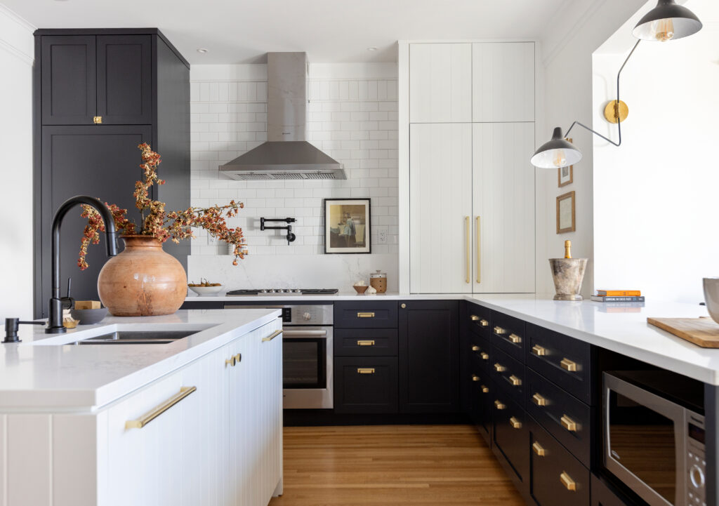 Modern kitchen with black and white cabinets, brass handles, white countertops, a large ceramic vase with branches on the island, stainless steel appliances, wood floor, and wall art.