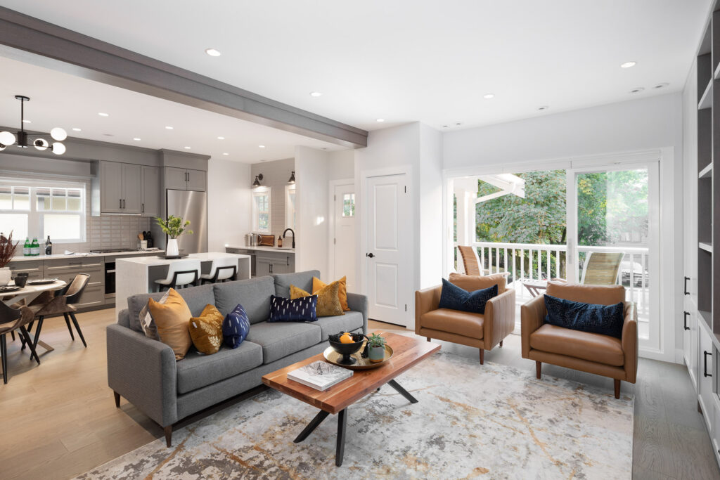 Modern open-concept living room and kitchen with gray sofa, brown armchairs, wood coffee table, and sliding glass doors leading to a patio. Neutral tones with colorful pillows and decorative accents.
