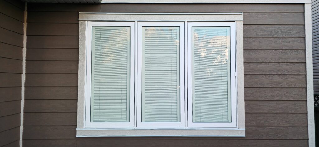 A modern house exterior with three white-framed windows, each covered by closed blinds, set in brown horizontal siding.