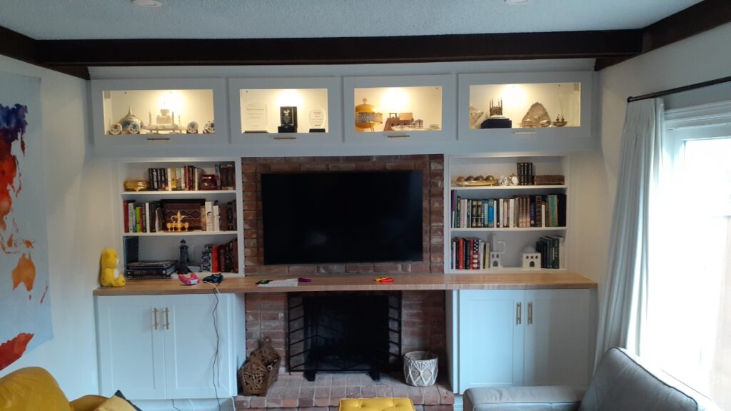 Built-in living room shelving with display cases above, featuring lit collectibles and awards. A flat-screen TV is set in the center above a brick fireplace, with bookshelves and cabinets on either side.