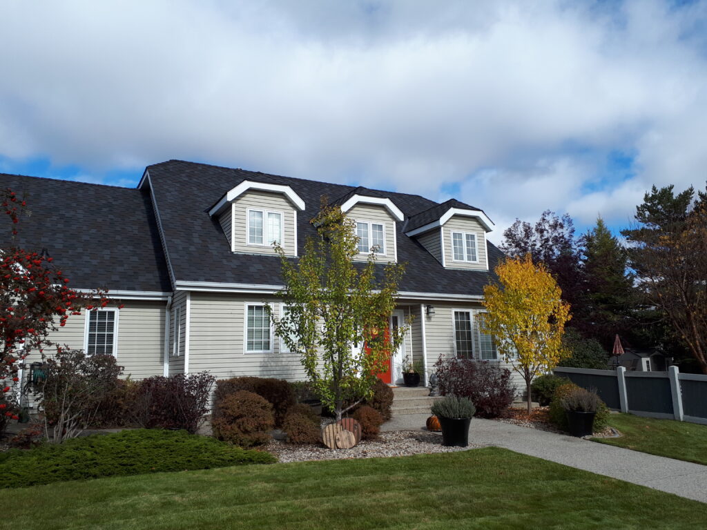 A beige two-story house with dormer windows and a dark roof sits behind a landscaped front yard with trees, shrubs, and a paved walkway under a partly cloudy sky.