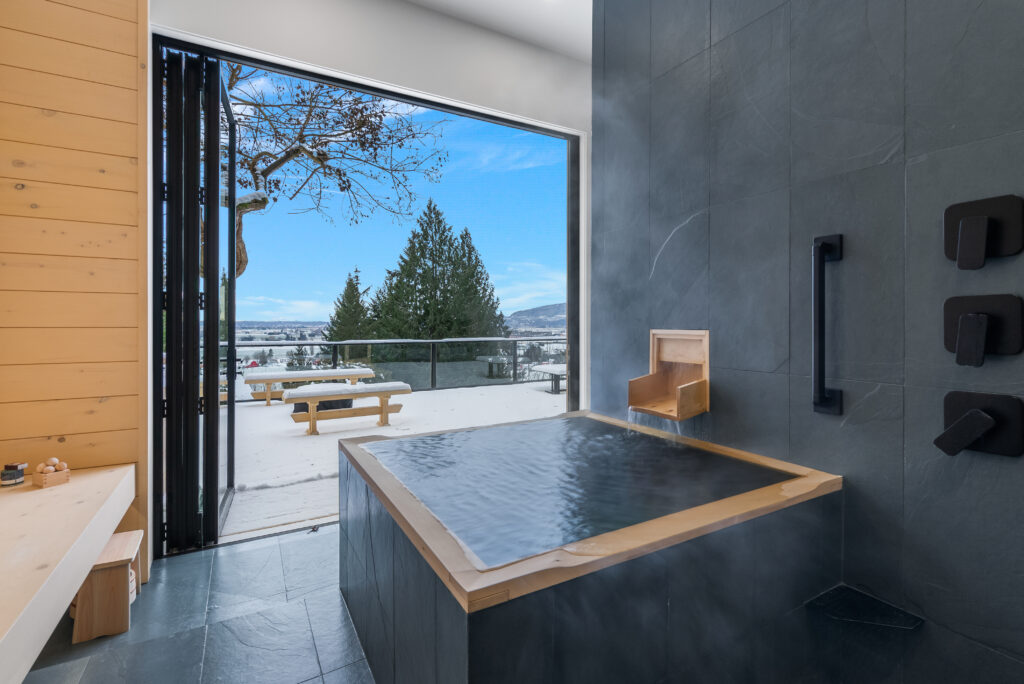 A modern indoor soaking tub with steam rising, open to an outdoor snowy landscape with a picnic table, trees, and distant mountains under a clear blue sky.