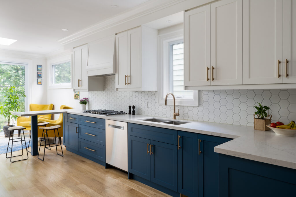 Modern kitchen with white upper cabinets, blue lower cabinets, hexagon tile backsplash, stainless steel appliances, and a dining area with yellow chairs by a window. Light wood floors and natural light brighten the space.