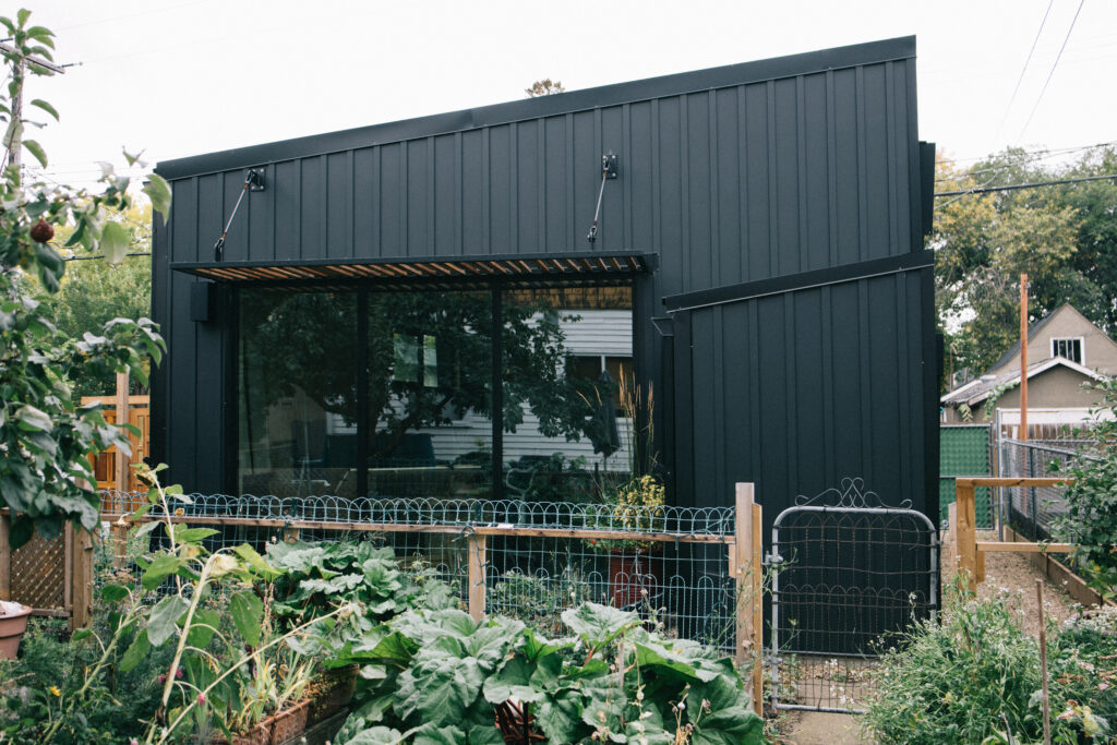 A modern black house with large glass windows is surrounded by a lush garden and enclosed by a wire fence and gate. Trees and neighboring houses are visible in the background.