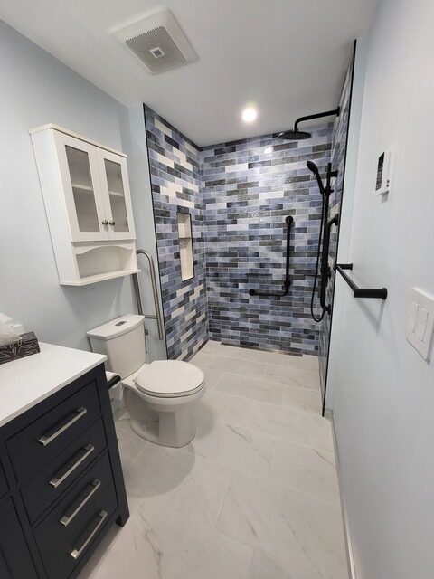 Modern accessible bathroom with blue and white tiled walk-in shower, black grab bars, wall-mounted showerhead, white toilet, navy vanity with drawers, and white wall cabinet. Light gray floor tiles.