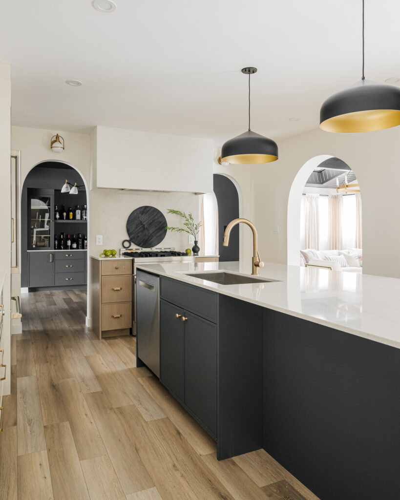 Modern kitchen with black and wood cabinets, white countertops, gold accents, two black pendant lights, and light wood floors. Arched doorways lead to dining and living areas with natural light.
