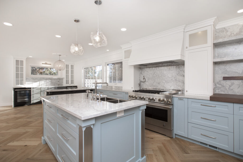 Modern kitchen with light blue cabinets, white marble countertops, a stainless steel oven, a large island with a sink, glass pendant lights, wooden herringbone floor, and large windows bringing in natural light.