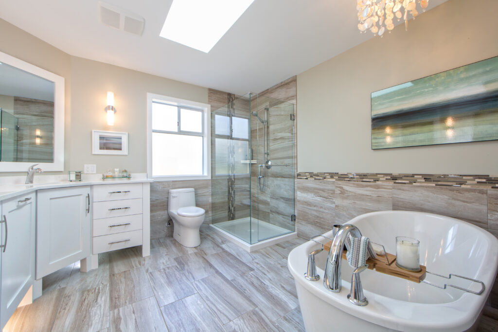 Modern bathroom with a white freestanding tub, glass-enclosed shower, toilet, and vanity. Light wood floors, neutral walls, wall art, and a skylight create a bright, airy atmosphere.