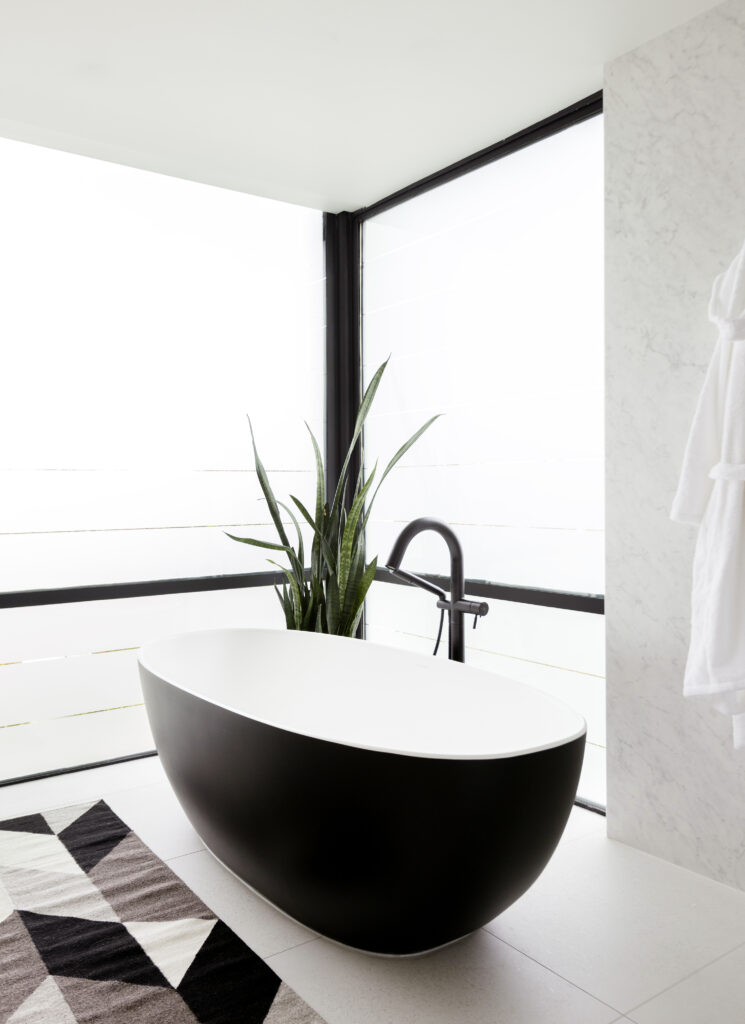 A modern bathroom with a black and white freestanding tub, a tall green plant beside it, a geometric-patterned rug, and large frosted windows letting in natural light.