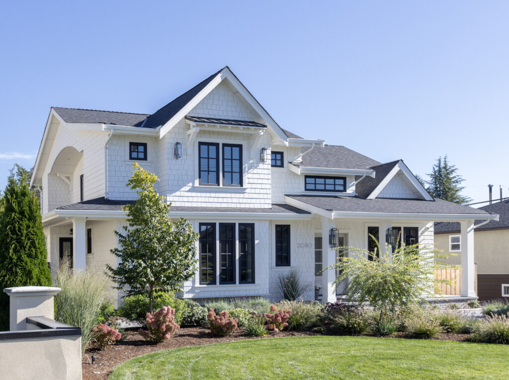 A modern two-story white house with black window frames, surrounded by green grass, trees, and landscaped shrubs under a clear blue sky.