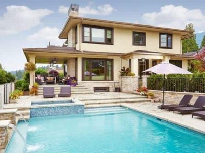 A modern two-story house with large windows, a patio area, and an outdoor pool surrounded by lounge chairs, plants, and a sun umbrella under a partly cloudy sky.