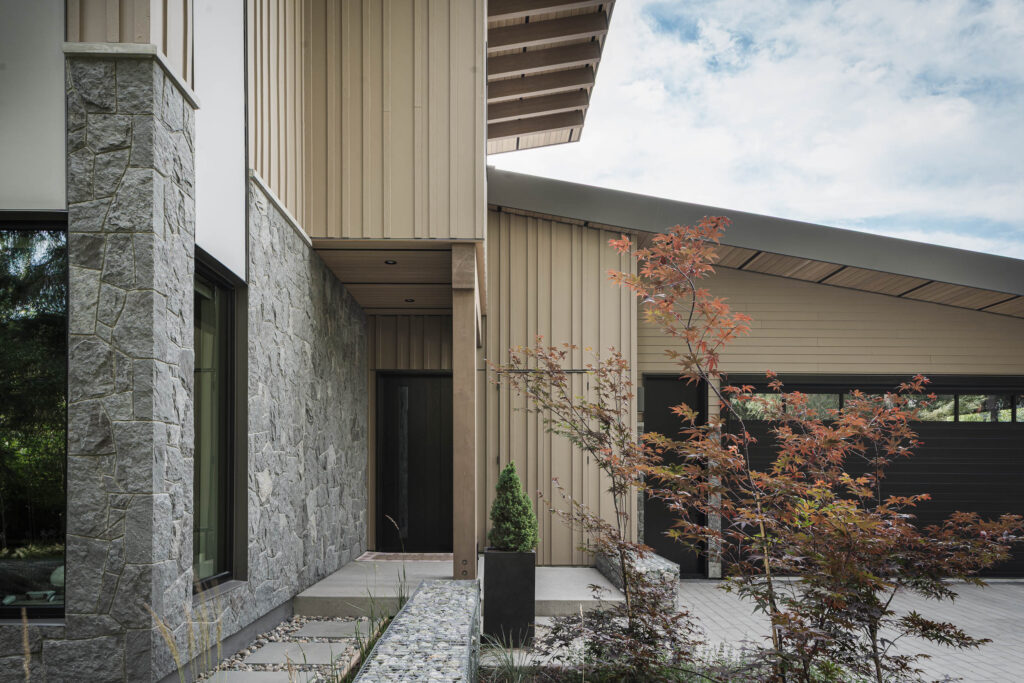 Modern house exterior with light wood paneling, stone walls, large dark-framed windows, and a black garage door. A small tree and landscaped garden are in the foreground under a partly cloudy sky.