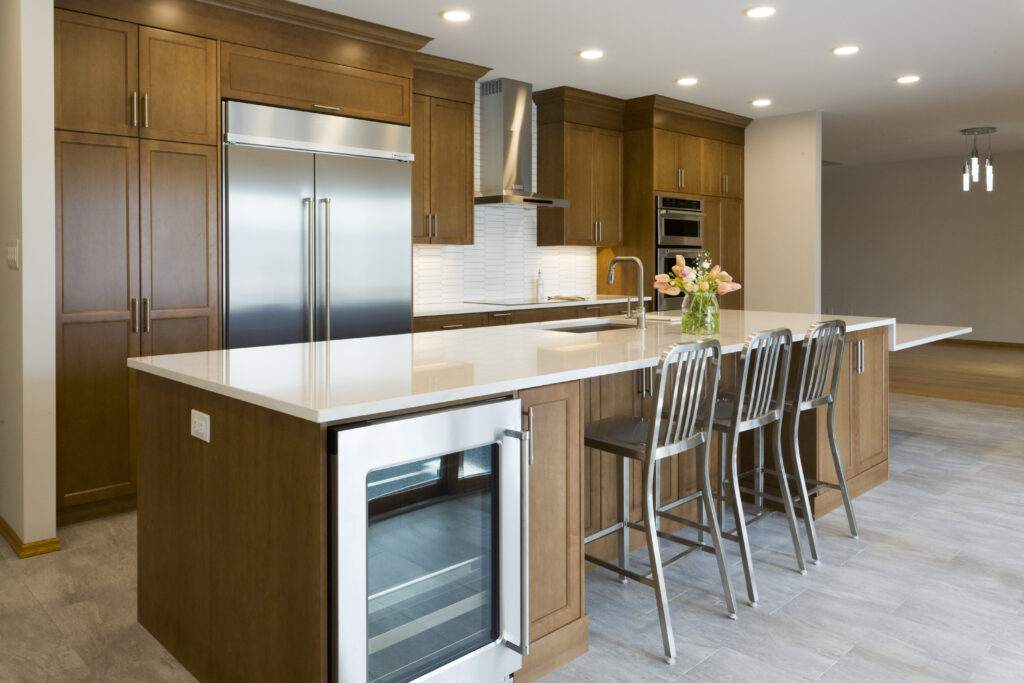 Modern kitchen with wood cabinets, stainless steel appliances, a large island with a white countertop, three metal barstools, a sink, and a vase of flowers. The space is bright with recessed lighting.