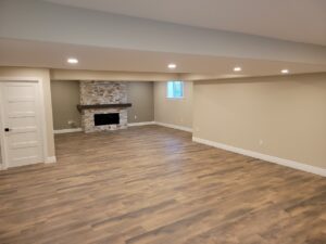 A spacious, empty basement room with light wood flooring, beige walls, recessed ceiling lights, a white door, and a stone fireplace with a small window above it.