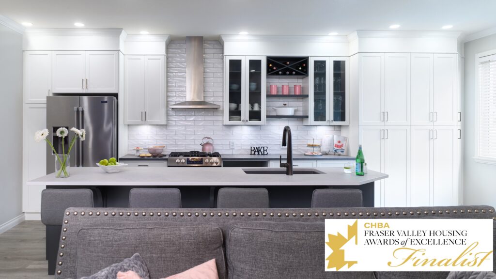 Modern white kitchen with stainless steel appliances, a large island with gray chairs, glass cabinets displaying dishes, and a Fraser Valley Housing Awards of Excellence Finalist badge in the lower right corner.