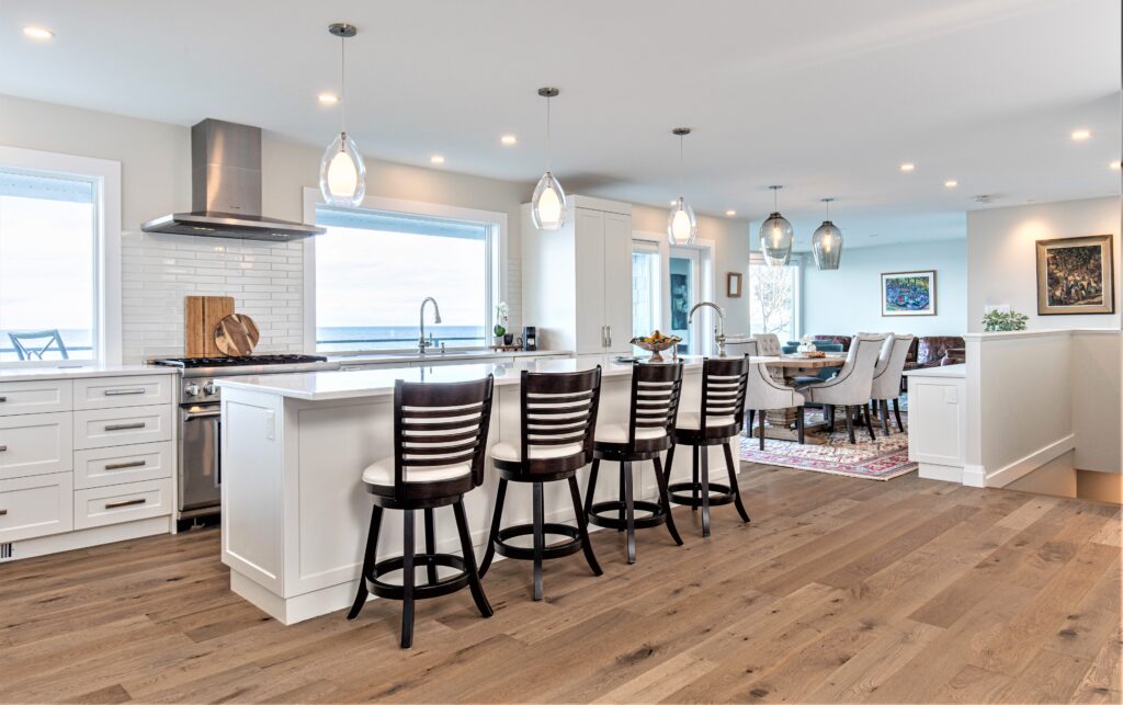 Modern open-concept kitchen with a large island, four dark-wood barstools, pendant lighting, stainless steel appliances, white cabinets, and hardwood floors. Dining area and living space are visible in the background.