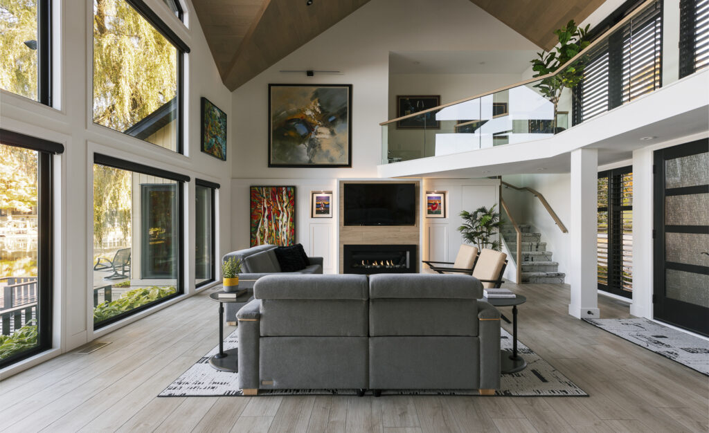A modern living room with large windows, gray sofas, a fireplace, colorful artwork on white walls, and an open staircase leading to an upper-level balcony. Light wood floors and abundant natural light fill the space.