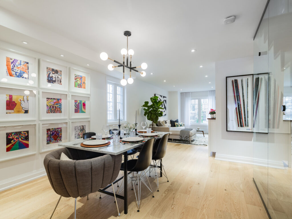 A modern dining and living area with light wood floors, a marble dining table set for four, contemporary chairs, colorful artwork on the walls, a large plant, and a cozy sitting area near windows.