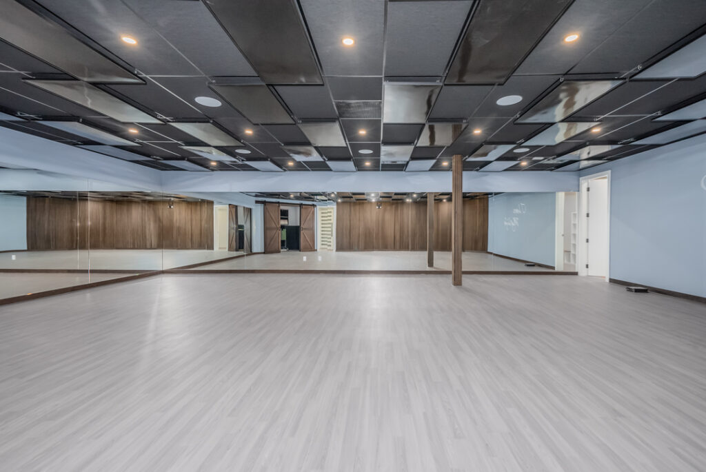 Spacious room with light gray flooring, wood-paneled walls, a mirrored wall, black-accented ceiling panels, recessed lighting, and an open doorway on the right. The room is empty and modern.