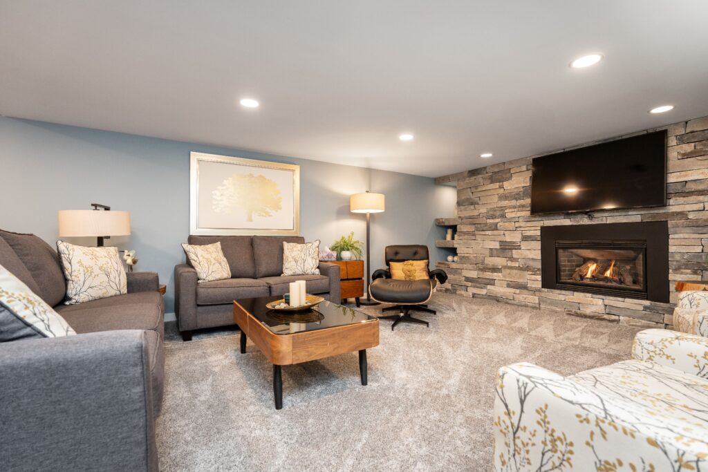 A cozy living room with gray sofas, a patterned armchair, a wood coffee table, a modern lounge chair, a stone fireplace with a TV above it, and soft lighting. The walls are light blue, and the carpet is light gray.