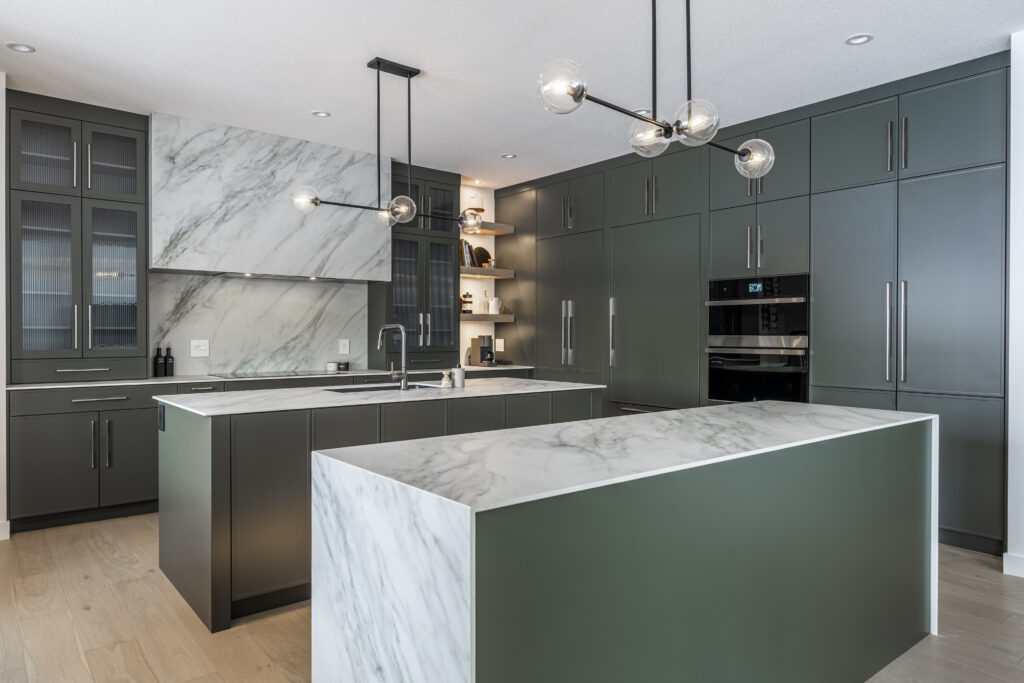 Modern kitchen with dark green cabinets, marble countertops and backsplash, built-in appliances, and two contemporary pendant lights hanging over a central island and a peninsula. Light wood floor and open shelving are also visible.