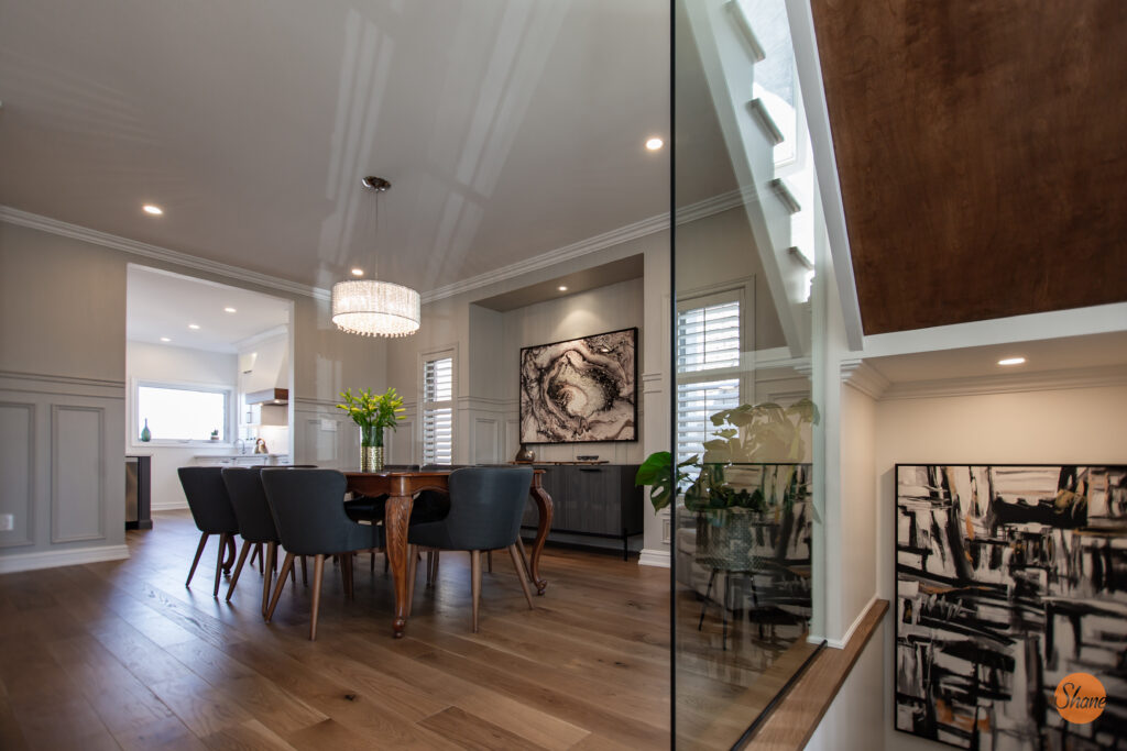 A modern dining room with a wooden table, six dark chairs, a glass chandelier, abstract artwork on the walls, and hardwood floors. Bright, natural light streams in from windows and an adjacent kitchen is visible in the background.