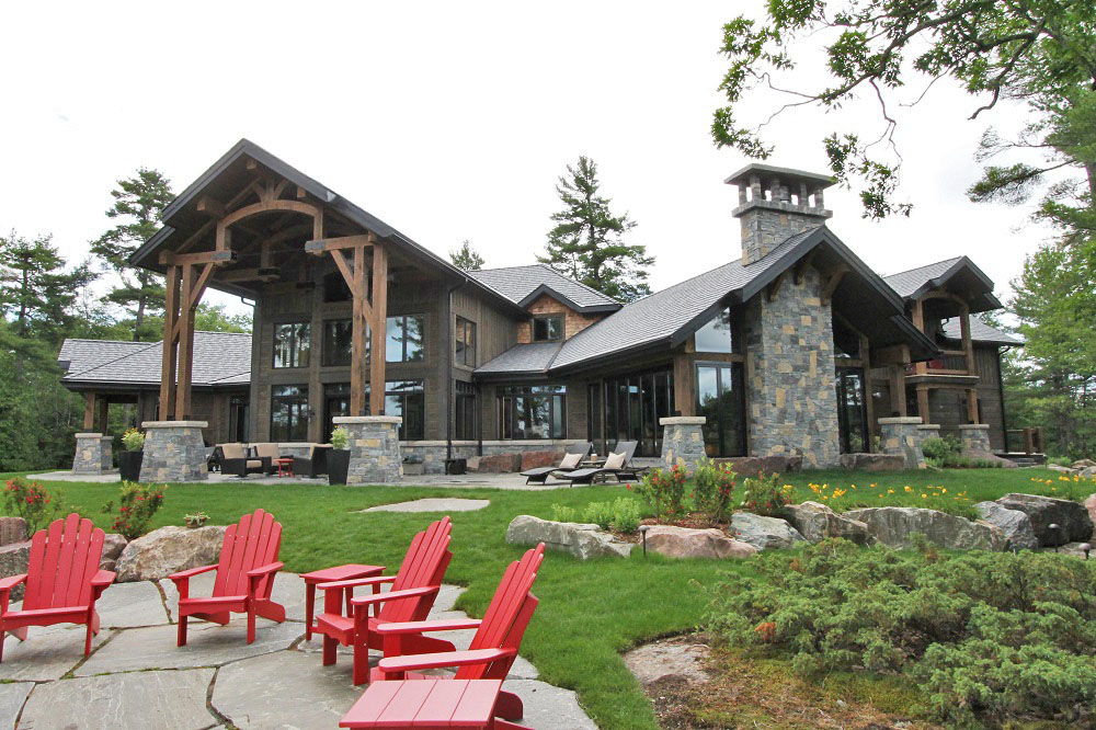Large rustic house with stone and wood exterior, many windows, and a spacious patio. Red Adirondack chairs are arranged around a stone fire pit on a stone patio, surrounded by green grass and trees.