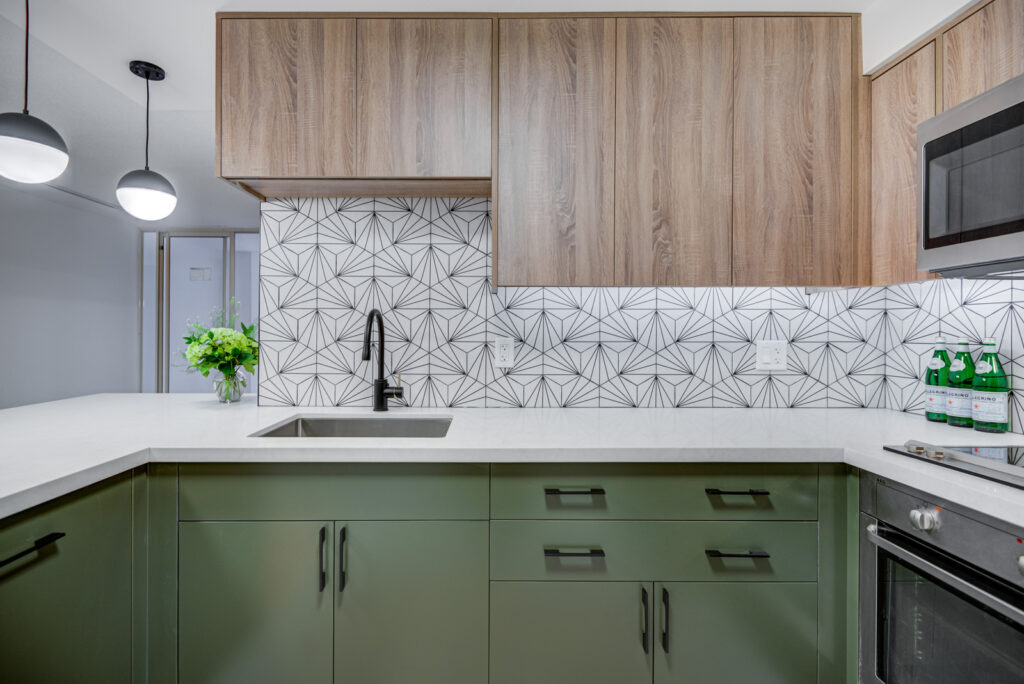 Modern kitchen with green lower cabinets, wood upper cabinets, geometric white and black tile backsplash, black faucet, white countertop, built-in stove, microwave, and a vase of green flowers. Two pendant lights hang above.