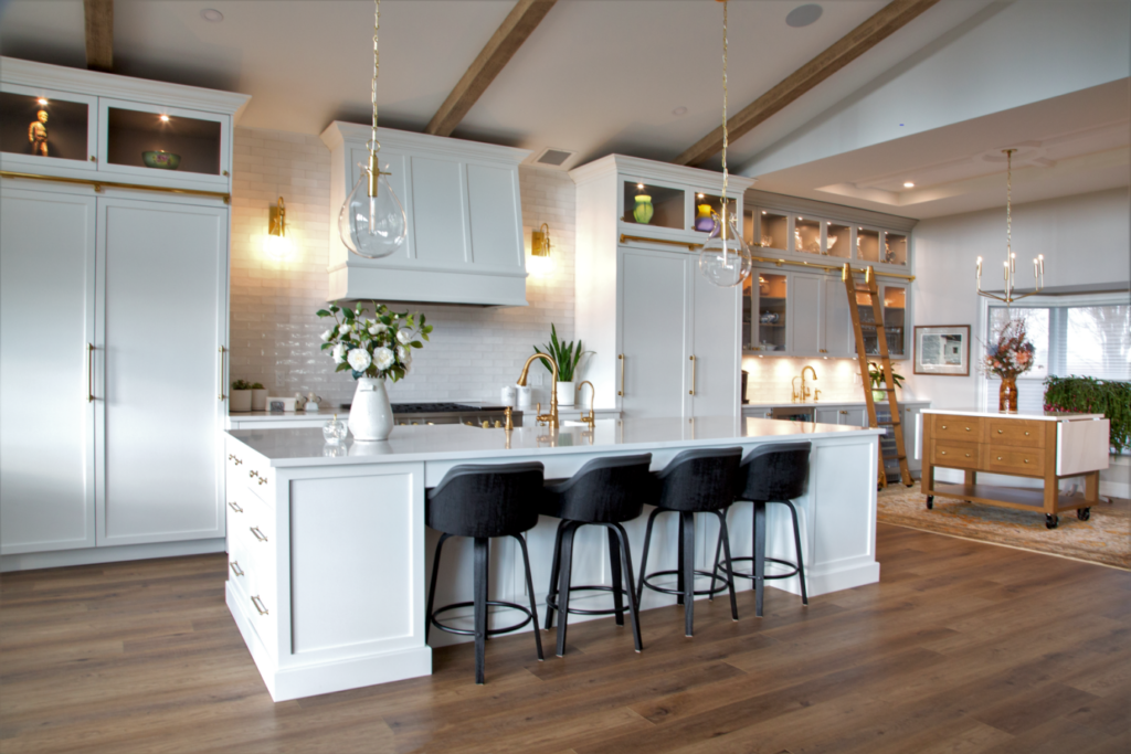 Modern kitchen with white cabinets, a large island with a marble countertop, three black barstools, pendant lights, wood flooring, and gold accents. There are flowers and decor on the island, and an open dining area in the background.