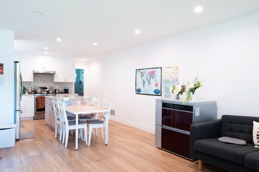 A bright, modern kitchen and dining area with white cabinets, a wooden floor, a dining table with white chairs, a world map on the wall, and a black sofa with a cushion. Flowers sit on a silver appliance.