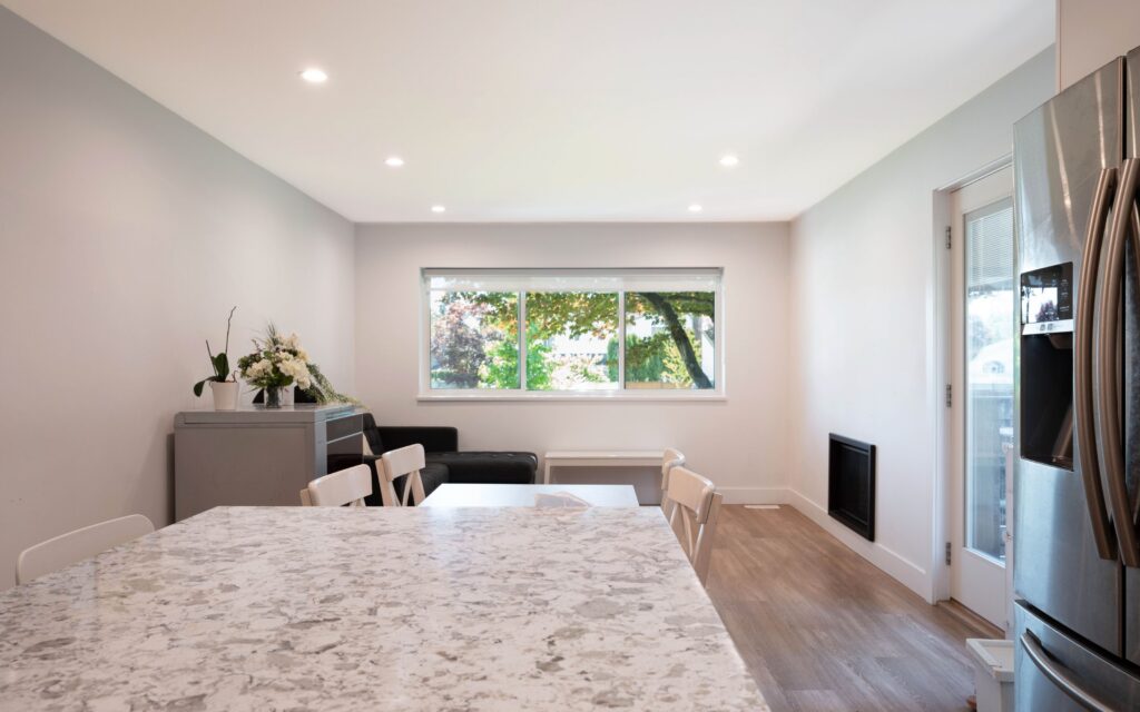 A modern, bright kitchen and dining area features a marble countertop, white chairs, light wood floors, a stainless steel refrigerator, and a large window overlooking greenery. A small flower arrangement sits on a sideboard.