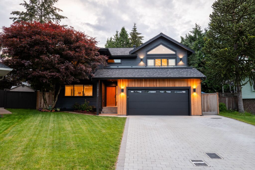 Modern two-story house with dark siding, large front windows, and a double garage. The home features warm exterior lights, a neatly mowed lawn, a paved driveway, and a large tree to the left.
