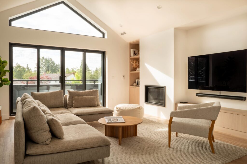 A modern living room with large windows, a grey sectional sofa, a beige armchair, a round wooden coffee table, a wall-mounted TV, a fireplace, built-in shelves, and light wood flooring. Natural light fills the space.